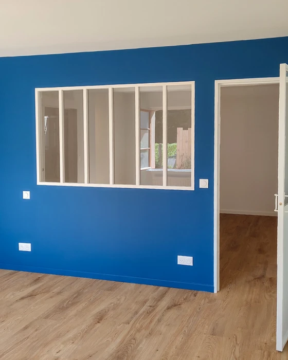 Photographie d'un intérieur en cours de renovation montrant un artisan applicant une couche de peinture sur un mur avec un pinceau ou rouleau, vêtu de manière professionnelle, dans un contexte résidentiel clair. La scène doit montrer le process de travail de peinture intérieure en action, avec des éléments visibles comme un pot de peinture, du matériel professionnel, et un mur partiellement peint pour illustrer l'avant/après ou le chantier en cours.